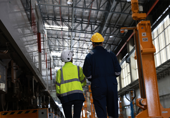Two health and safety workers in hard helmets and hi-vis jackets walking around a UK warehouse
