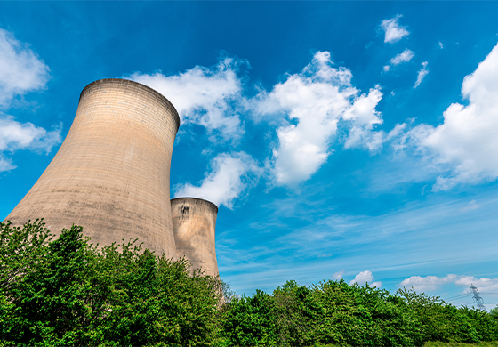 Two cooling towers standing behind a hedgerow with a sunny sky above.