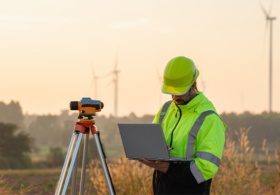 A health and safety man in a hi-vis and helmet looking at a laptop while using a measuring device in a UK field surrounded by wind turbines.